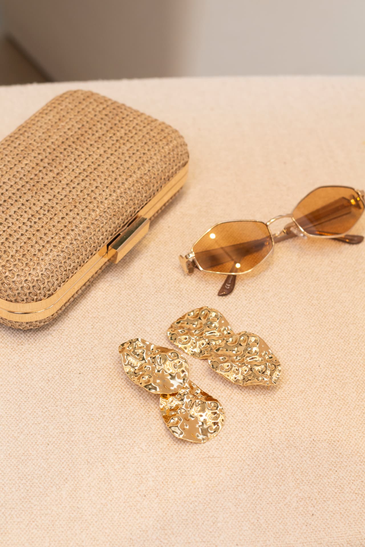 Brincos dourados texturizados ao lado de clutch e óculos de sol